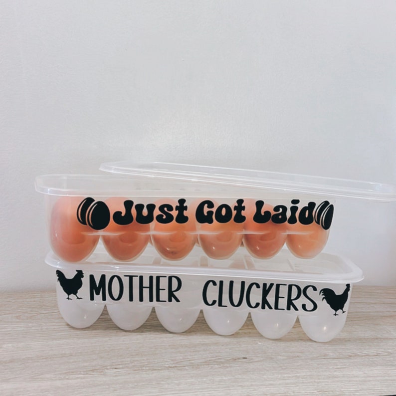 May include: Two clear plastic egg containers, one stacked on top of the other. The top container has the text "Just Got Laid" in black. The bottom container has the text "Mother Cluckers" and a silhouette of a chicken.