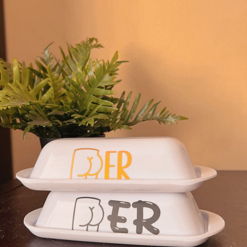 May include: Two white ceramic butter dishes stacked on top of each other. The top dish has a gold graphic of a rear end and the letters "PER". The bottom dish has a gray graphic and letters. A green plant is in the background.