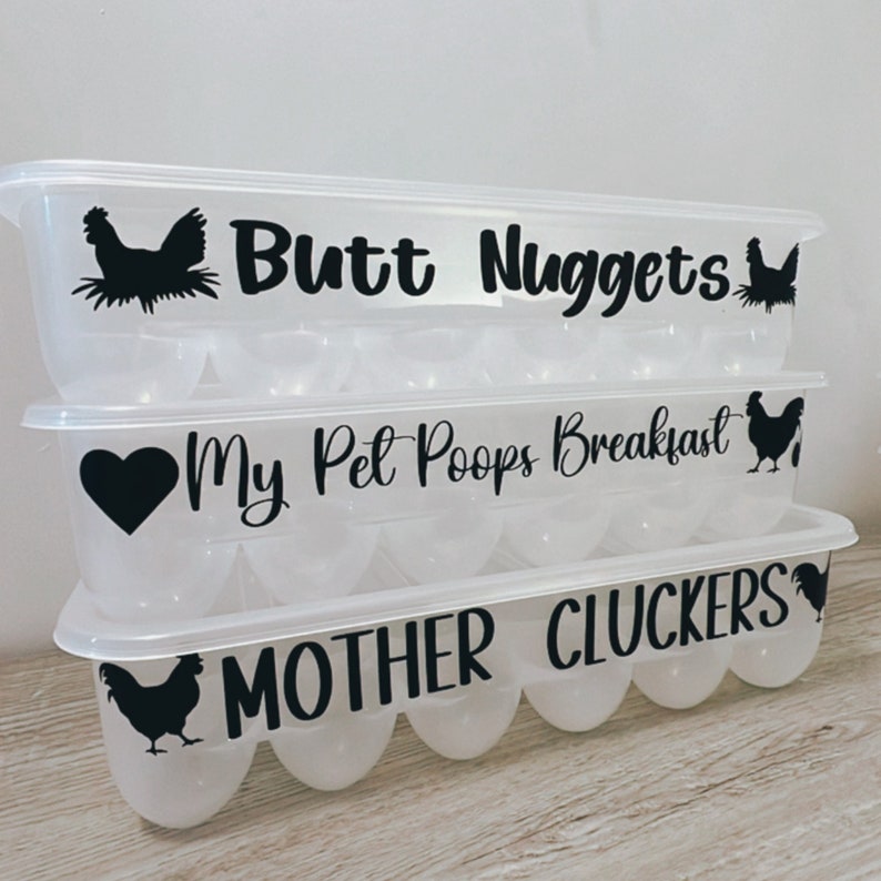 May include: Three clear plastic egg containers stacked on top of each other. Each container has a black graphic and text: "Butt Nuggets", "My Pet Poops Breakfast", and "MOTHER CLUCKERS". The containers are designed to hold eggs.