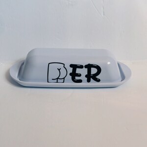 May include: A light blue butter dish with a lid and a tray. The dish has a black graphic of a butt and the letters "PER" printed on the front. The dish is on a white surface.