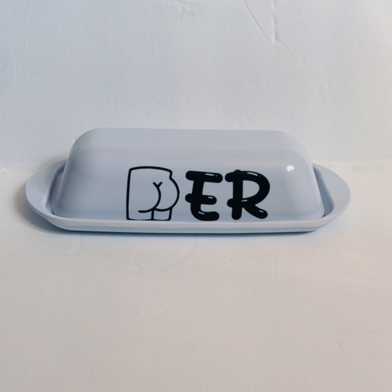 May include: A light blue butter dish with a lid and a tray. The dish has a black graphic of a butt and the letters "PER" printed on the front. The dish is on a white surface.