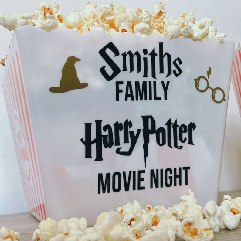 May include: White popcorn bucket with red and white stripes. The bucket has a black and white graphic that says "Smiths Family Harry Potter Movie Night". There is a black silhouette of a witch's hat and a pair of glasses.