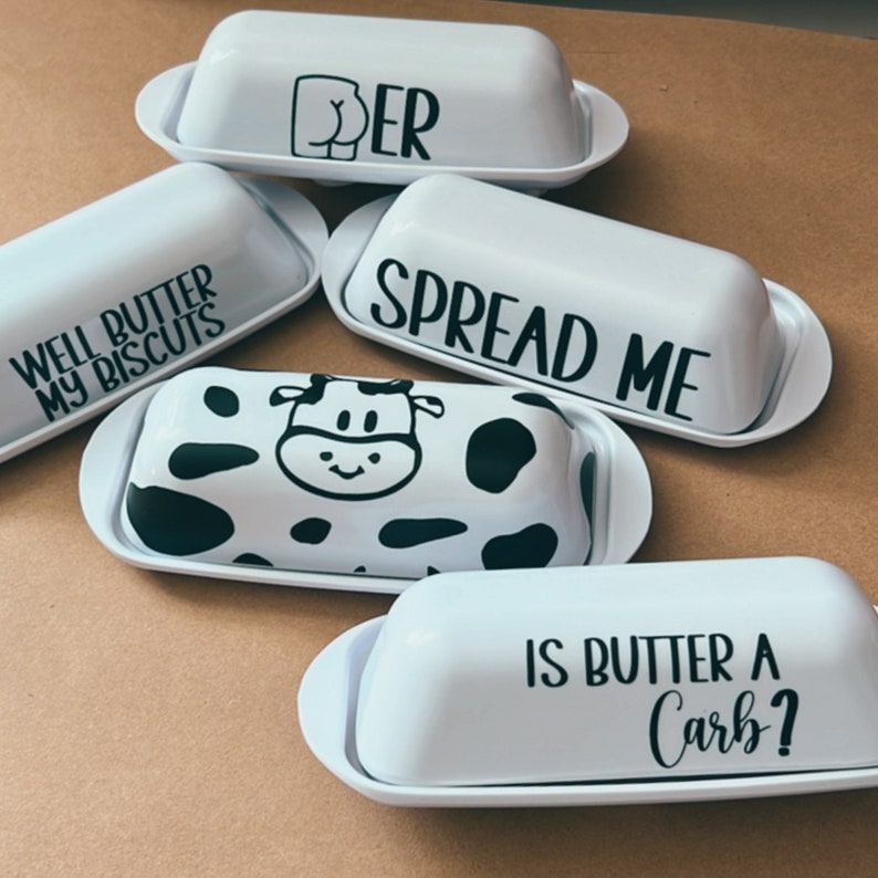 May include: A collection of white ceramic butter dishes with various black graphic designs and text. The designs include a cow pattern, the word "SPREAD ME", the phrase "WELL BUTTER MY BISCUITS", the phrase "IS BUTTER A Carb?", and a graphic of a rear end.