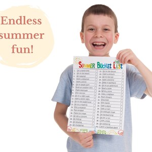 May include: A young boy smiles while holding a summer bucket list with 42 activities to complete. The list includes activities like going to the beach, having a picnic, visiting the zoo, and reading a book.