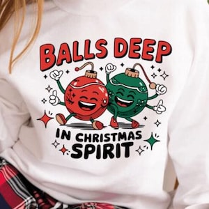 May include: White sweatshirt with a graphic that reads "BALLS DEEP IN CHRISTMAS SPIRIT." The graphic features two cartoon Christmas ornaments, one red and one green, with smiling faces and arms raised in celebration. The design includes stars and decorative elements.