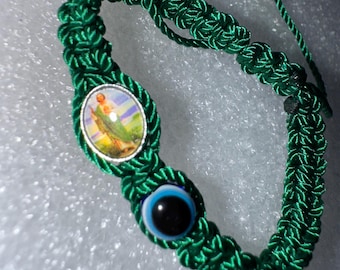 May include: A green macrame bracelet featuring an oval-shaped religious medallion and a blue evil eye bead. The bracelet is made of woven green cord and has an adjustable sliding knot closure.
