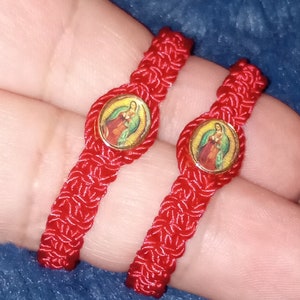 May include: Two red string bracelets with a gold-colored charm featuring a religious image. The charms are round and have a colorful image of a woman and child.