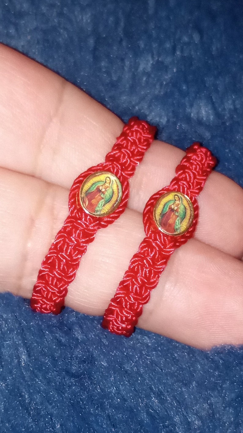 May include: Two red string bracelets with a gold-colored charm featuring a religious image. The charms are round and have a colorful image of a woman and child.