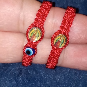 May include: Two red string bracelets with charms. One bracelet has a blue evil eye charm and the other has a gold charm with a religious image.