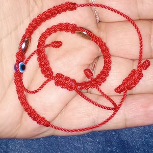 May include: Red string bracelet with a blue evil eye charm and silver beads. The bracelet is knotted and tied together.