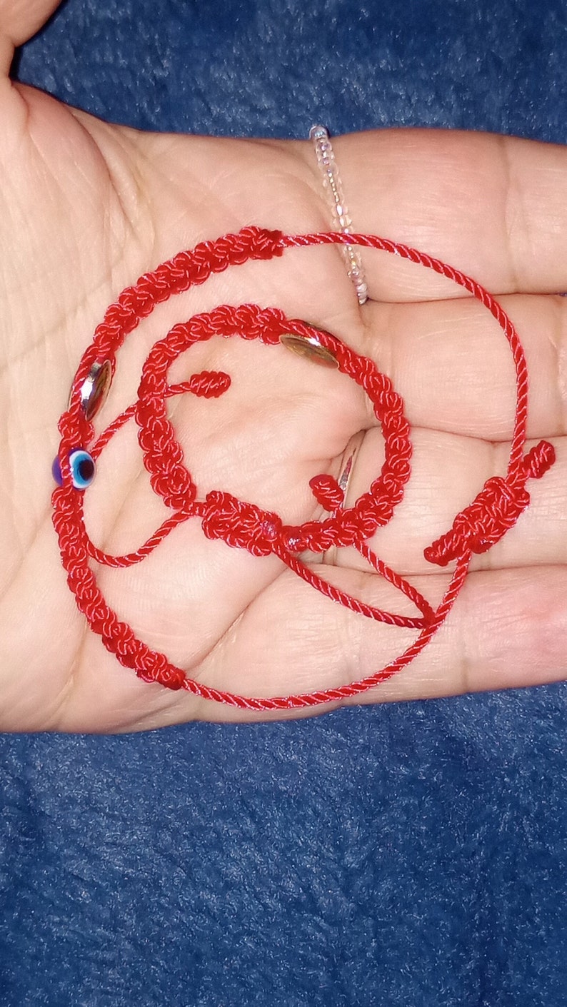 May include: Red string bracelet with a blue evil eye charm and silver beads. The bracelet is knotted and tied together.