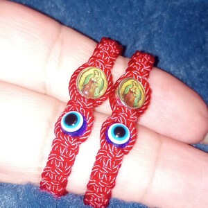 May include: Two red string bracelets with blue evil eye charms and a religious image charm. The bracelets are tied together and are on a finger.