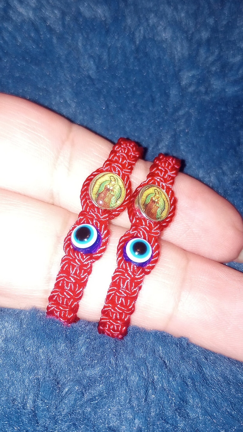 May include: Two red string bracelets with blue evil eye charms and a religious image charm. The bracelets are tied together and are on a finger.