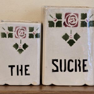 Può includere: Due contenitori rettangolari in ceramica, uno con la scritta "THE" e l'altro "SUCRE" in lettere nere. Entrambi hanno uno sfondo bianco con un motivo floreale decorativo in verde e rosso. I contenitori sono probabilmente per la conservazione in cucina.