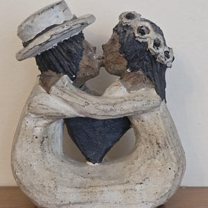 May include: A ceramic sculpture of a couple embracing and kissing. The figures have dark hair and skin, with the man wearing a hat. The sculpture is primarily off-white with black accents, and a black heart shape is visible.