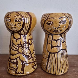 May include: Two ceramic vases with a tan glaze and brown line drawings. The vases are shaped like figures with stylized faces and long hair. One vase depicts a figure holding a child. The other vase depicts a figure with arms crossed.