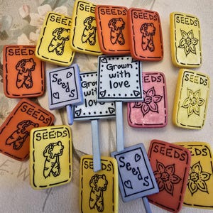 May include: A selection of decorative seed packet-shaped items in various colours. The packets are yellow, orange, pink, and red, with the word "SEEDS" and a carrot or flower graphic. Some have small signs that say "Grown with love".