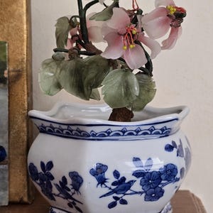 Puede incluir: Un bonsái decorativo con flores rosas y hojas verdes, en una maceta de porcelana blanca y azul. La maceta presenta un diseño floral y de pájaros azul. Un artículo de decoración del hogar único.