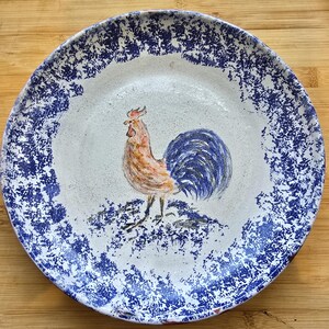 May include: A white ceramic plate with a blue speckled border and a hand-painted rooster in the centre. The rooster has orange, brown, and blue feathers. The plate is on a wooden surface.