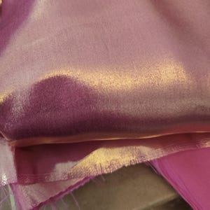 May include: A folded piece of iridescent pink fabric with a smooth, reflective surface that shifts between light and dark pink tones. The material has a subtle sheen and visible loose threads along the raw, unfinished bottom edge of the textile.