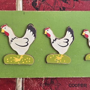Può includere: Un puzzle in legno vintage con cinque galline bianche con accenti neri e rossi, ciascuna in piedi su una base verde con dettagli gialli. Le galline sono disposte in ordine decrescente di dimensioni su una tavola rettangolare verde.