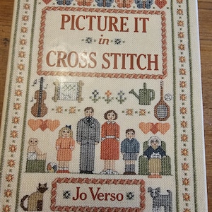 May include: A book cover with a cross stitch design of a family, a cat, a dog, and a garden scene. The title of the book is "Picture It in Cross Stitch" by Jo Verso.
