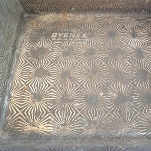 Può includere: Una teglia quadrata in metallo con un motivo a raggiera testurizzato sulla superficie interna. Il centro presenta il testo in rilievo OVENEX, RD 861592, PAT 495306 e MADE IN ENGLAND. Il metallo mostra segni di invecchiamento con ossidazione.