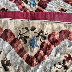 May include: Close-up of a vintage textile with a repeating pattern. The design features alternating rows of cream, red, and white geometric shapes. Small blue floral accents and a red ribbon add colour and detail to the textured fabric.