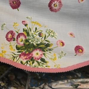 Könnte beinhalten: Ein hellblauer Stoff mit einem floralen Druckmuster aus rosa und weißen Gänseblümchen mit grünem Blattwerk. Die geschwungene Kante des Stoffes ist mit einer strukturierten rosa Borte versehen. Das Material hat ein sichtbares Gewebe.