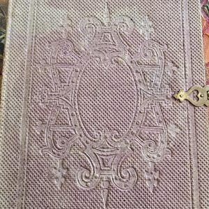 Puede incluir: Una cubierta de libro vintage, de color púrpura claro, con un diseño en relieve. La cubierta presenta un diseño central ornamentado en forma ovalada con la letra "A" incorporada. Un pequeño cierre metálico decorativo es visible en el lado derecho.