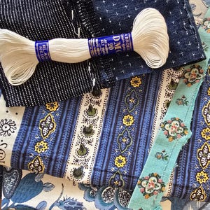 Op de afbeelding: Een assortiment van stofstalen in diverse patronen en kleuren, waaronder blauw-witte strepen, bloemmotieven en een textuurrijke marineblauwe stof. Een spoel wit borduurgaren met een blauw label ligt erbovenop.