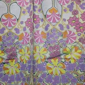 Op de afbeelding: Een stof met een herhalend patroon van roze, gele, oranje en paarse bloemen op een witte achtergrond. De bloemen zijn gestileerd en hebben een retro-gevoel uit de jaren 70.