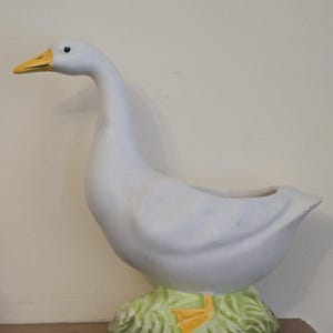 May include: A white ceramic goose planter with a yellow beak and feet. The planter has a green base with a grass design. The goose is standing upright with its neck curved. The planter is designed to hold plants or flowers.