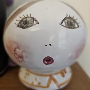 May include: A white ceramic figurine of a face with green eyes, a pink blush, and a red mouth. The figurine has a yellow collar and is standing on a wooden surface.