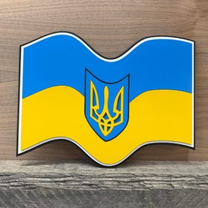 May include: A wooden sign featuring the Ukrainian flag. The flag design includes a blue top and yellow bottom, with a gold trident emblem in the center. The sign has a black border and a wavy shape.