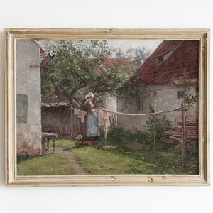May include: A framed oil painting depicting a woman hanging laundry in a backyard. The scene is set in front of a white building with a red roof. The woman is wearing a blue dress and a white apron. The painting is framed in a light wood frame.