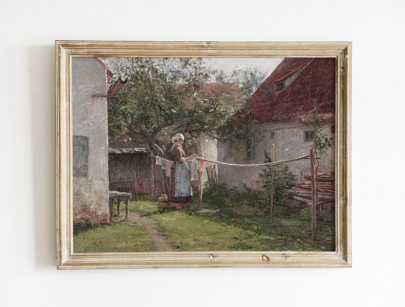 May include: A framed oil painting depicting a woman hanging laundry in a backyard. The scene is set in front of a white building with a red roof. The woman is wearing a blue dress and a white apron. The painting is framed in a light wood frame.