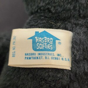 May include: Close-up of a plush toy with a tag that reads "HASBRO SOFTIES." The toy is dark gray with a red fabric accent. The tag also includes the Hasbro Industries, Inc. address in Pawtucket, RI.