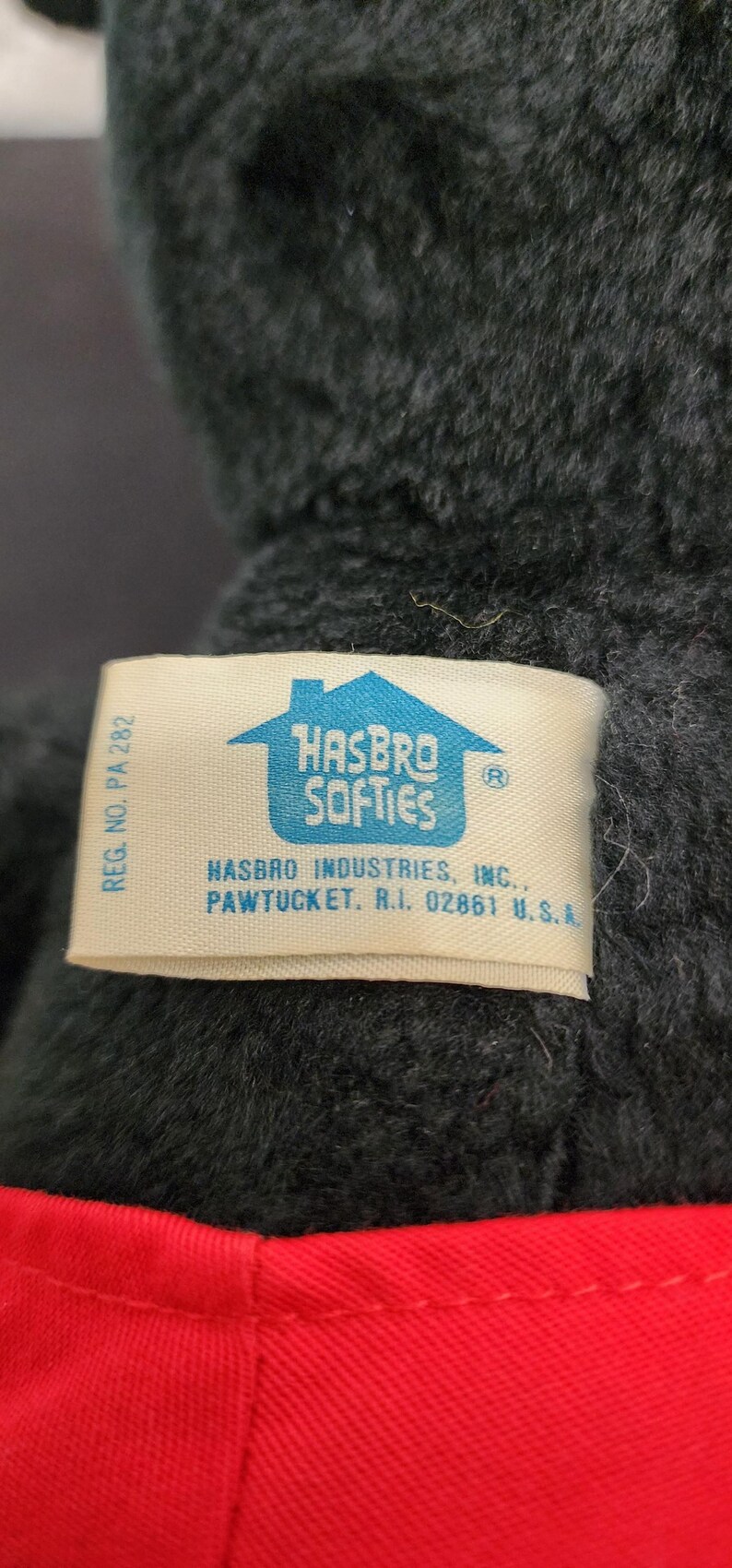 May include: Close-up of a plush toy with a tag that reads "HASBRO SOFTIES." The toy is dark gray with a red fabric accent. The tag also includes the Hasbro Industries, Inc. address in Pawtucket, RI.