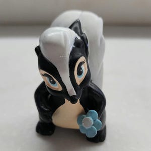 May include: A black and white skunk figurine with a blue flower. The skunk has a white stripe down its back and is holding a blue flower in its right paw.
