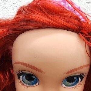 May include: A doll's face with red hair, blue eyes, and a pink bow. The doll's face is pale with painted eyebrows, eyelashes, and lips. The doll's hair is long and flowing, and the bow is on top of the head.