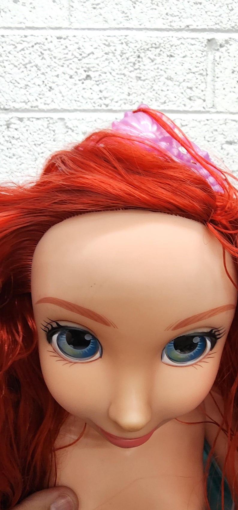 May include: A doll's face with red hair, blue eyes, and a pink bow. The doll's face is pale with painted eyebrows, eyelashes, and lips. The doll's hair is long and flowing, and the bow is on top of the head.