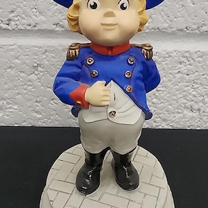 May include: A figurine of a young boy dressed as Napoleon Bonaparte. The figure wears a blue jacket with gold buttons, white pants, and black boots. The base is white with a brick pattern and a plaque that reads "Napoleon PLANT."