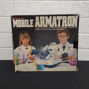 May include: Vintage Radio Shack Mobile Armatron box featuring a robot arm and two children in lab coats. The box has the text "MOBILE ARMATRON" and "A WIDE RANGE OF PLAY APPLICATIONS TO SATISFY YOUR EVERY DREAM OF CONTROLLING YOUR OWN ROBOT."
