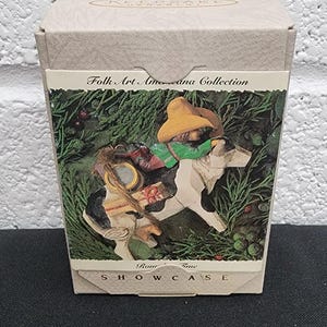 May include: A beige cardboard box for a Hallmark Keepsake Ornament from the Folk Art Americana Collection. The front features an illustration of a figure in a yellow hat riding a white horse, with the text Showcase and Rour Time visible.