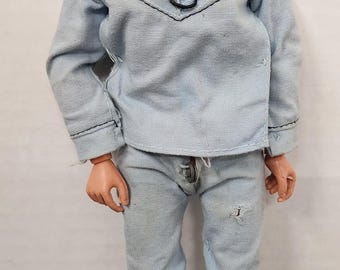 May include: A vintage action figure wearing a light blue shirt with a black lace-up neckline and matching light blue pants. The figure has brown hair and brown eyes.