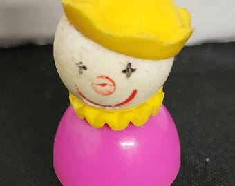 May include: A vintage plastic toy clown with a yellow hat, white face, red lips, and a pink body. The clown has black eyes with white stars.