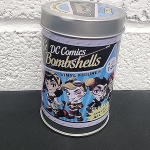 May include: A cylindrical tin featuring DC Comics Bombshells vinyl figures. The tin has a silver lid and a blue and purple design with images of the characters. The text "DC Comics Bombshells Vinyl Figures" is displayed prominently.