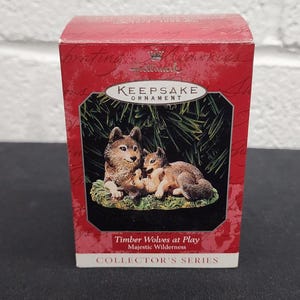 May include: A red and gold Keepsake Ornament box featuring a detailed image of two timber wolves. The box reads "Timber Wolves at Play" and "Collector's Series."