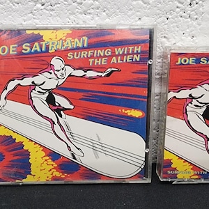 May include: Two music albums, a CD and a cassette tape, featuring the album art for Joe Satriani's "Surfing with the Alien." The artwork depicts a silver figure surfing on a silver board against a red, yellow, and blue background.
